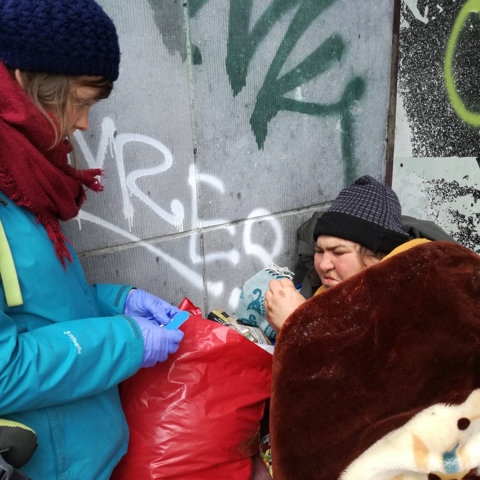 Cathy est dos à un mur rempli de graffitis. Elle se protège du froid grâce à un bonnet gris et une grosse couverture bordeaux. Autour d'elle se trouvent ses affaires. Elle est une personne sans-abri. © Infirmiersderue