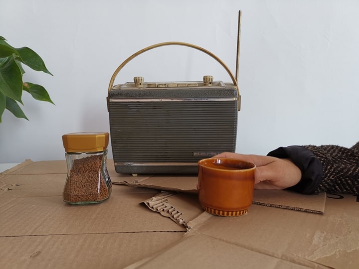 Une main est proche d'une tasse de café. Derrière se trouve un petit poste radio.